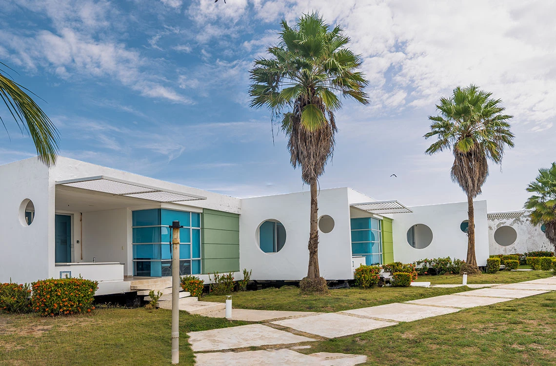Resort moderno con palmeras y Marina Puerto Velero. Habitaciones blancas con ventanas azules y verdes.