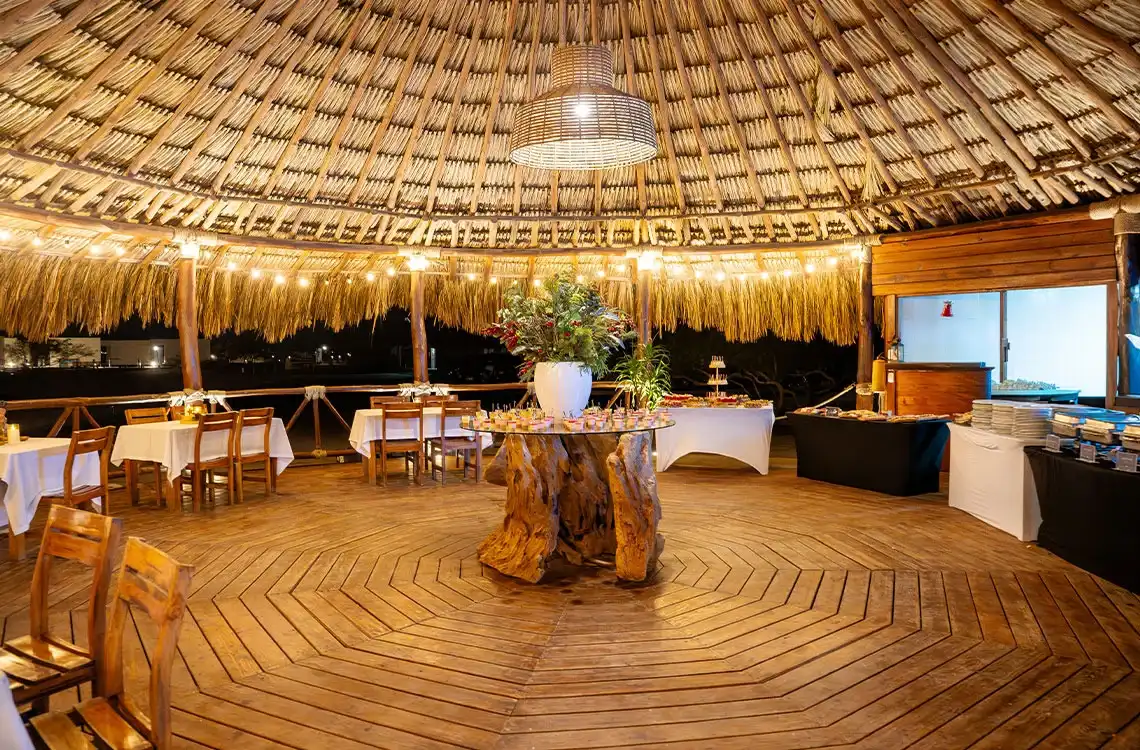 Salón tropical con techo de palapa, mesas para eventos y @Marina Puerto Velero@, ideal para bodas y conferencias.