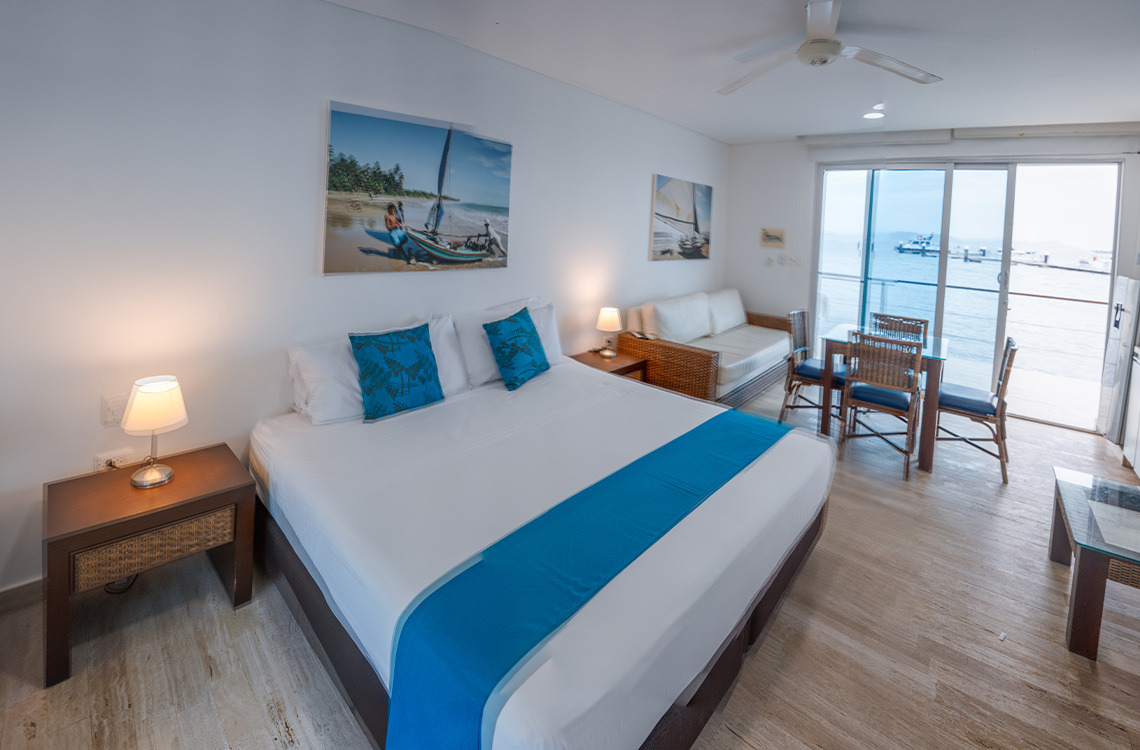 Amplia habitación de hotel con cama doble, sofás y mesa de comedor con vista al mar. Marina Puerto Velero