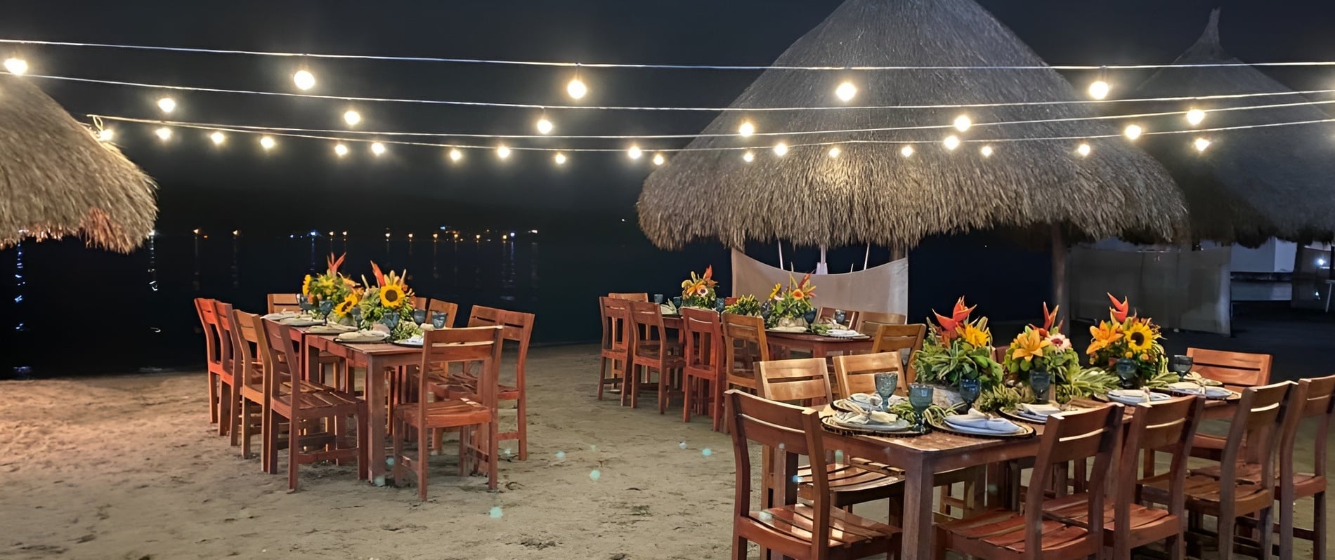 Mesas de cena en la playa bajo luces y palapas en Marina Puerto Velero.