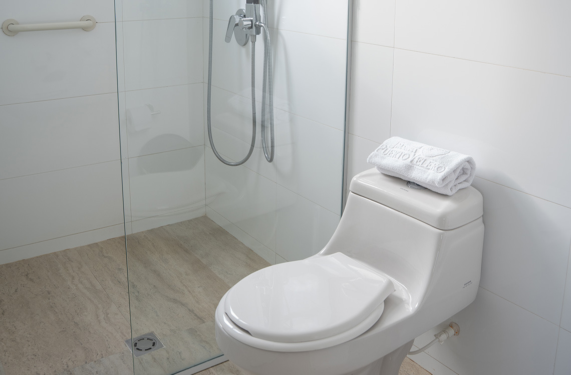 Baño moderno con ducha y WC, toalla blanca doblada con logo de Marina Puerto Velero.