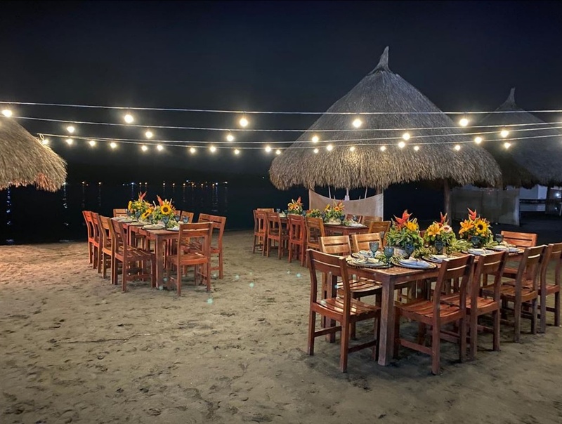Mesas de banquete en la playa, iluminadas por luces de feria, cerca de Marina Puerto Velero.