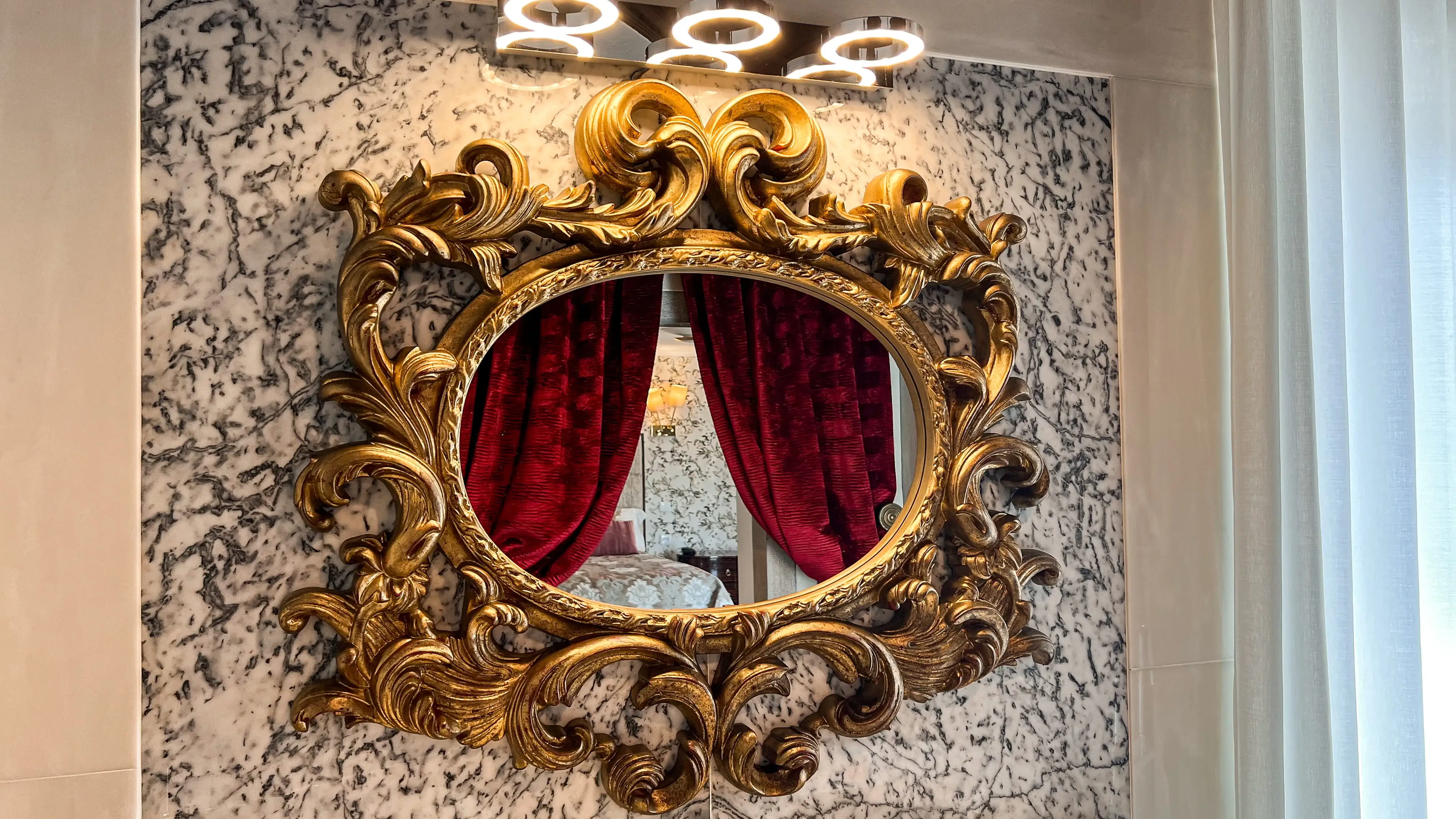 Espelho ornamentado em mármore refletindo cortinas de veludo e uma cama em hotel Marmoris Hotels.