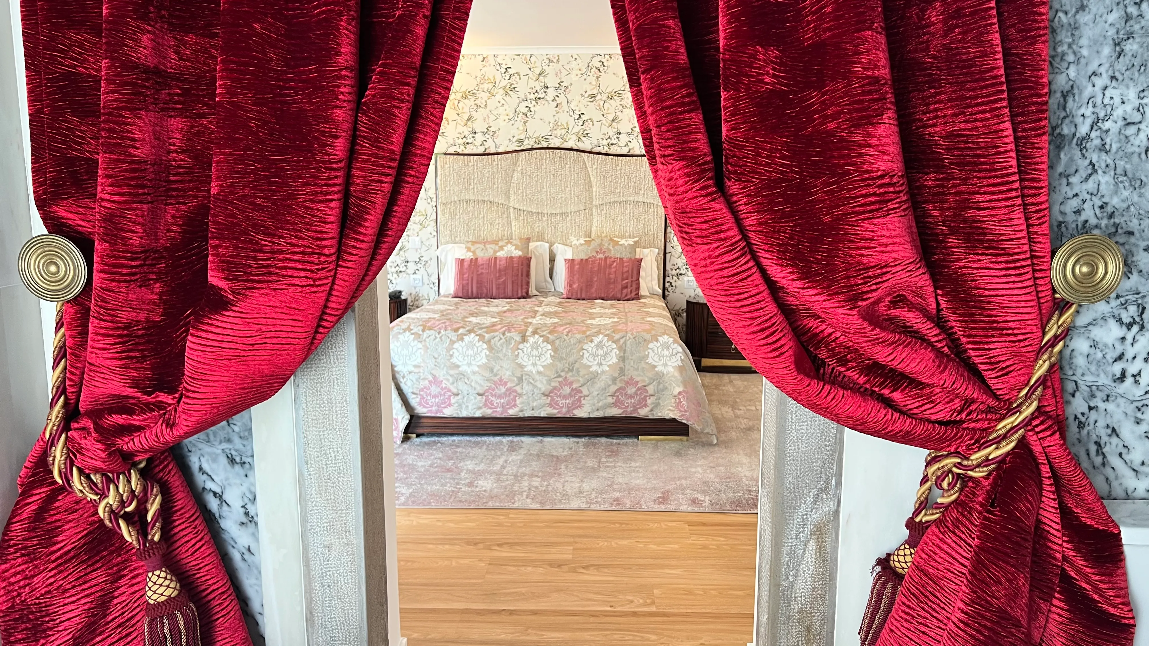 Cortinas vermelhas e luxuosas abrem vista para quarto de hotel com cama grande e papel de parede floral. Marmoris Hotels