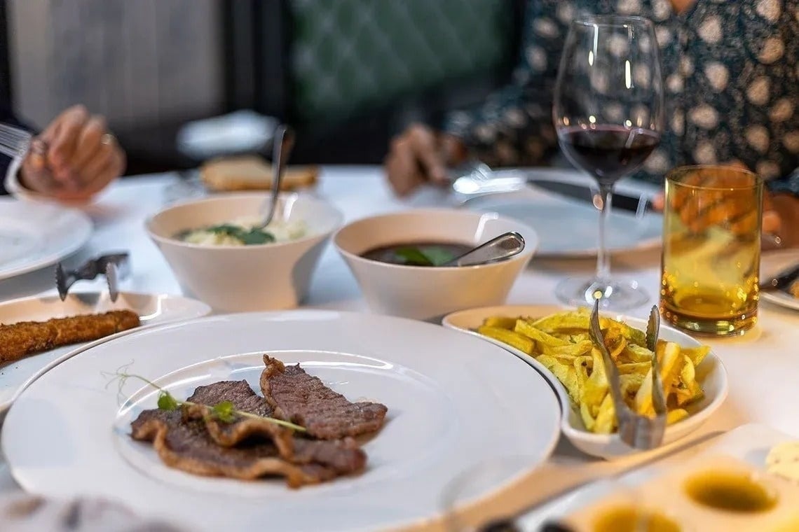 A dining table is laden with various dishes, including sliced meat, golden fries, and two bowls of soup, accompanied by red wine, with diners in the softly blurred background.