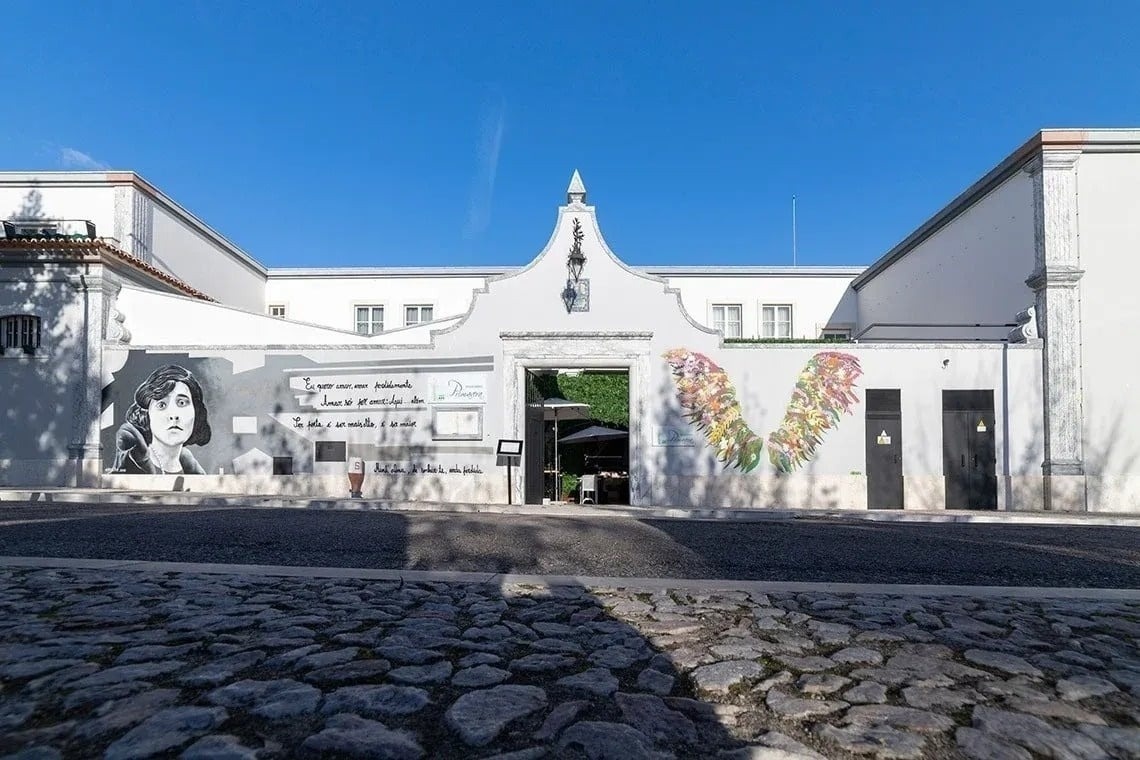 A white building with a prominent archway and murals, including a woman's portrait and colorful wings, is viewed from a low angle across a cobblestone street under a bright blue sky.