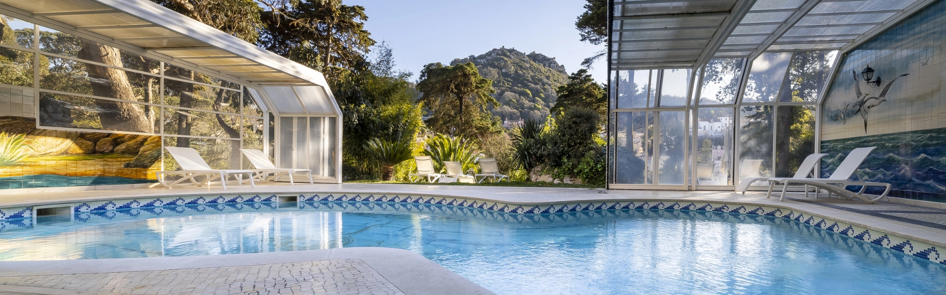 Indoor swimming pool with loungers and a mural Marmoris Hotels, surrounded by lush greenery and mountains.