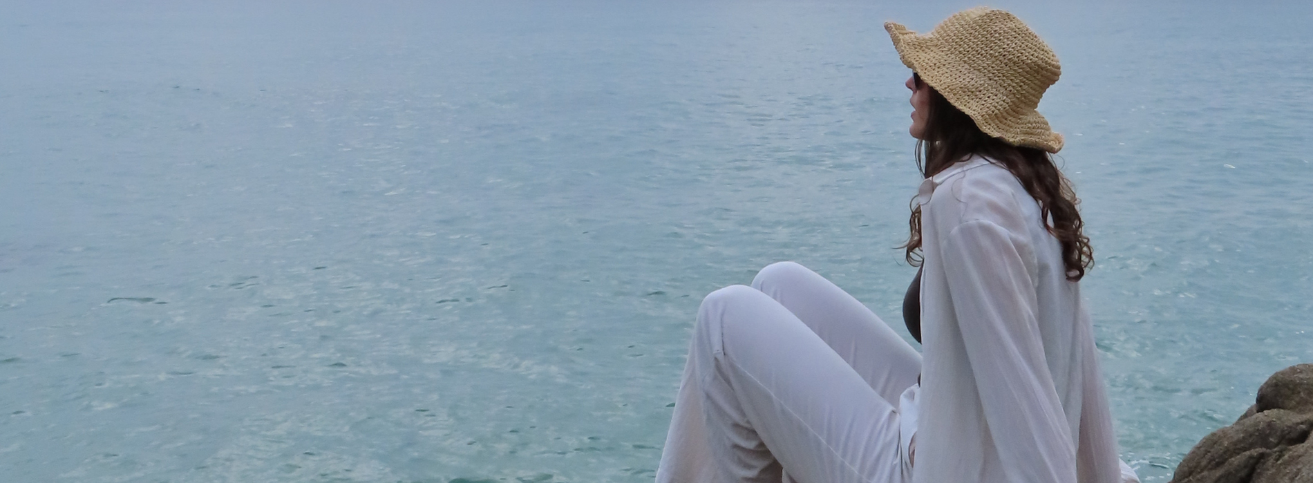 Una dona amb un barret de palla i vestida de blanc seu de costat a la vora del mar, contemplant l'aigua.