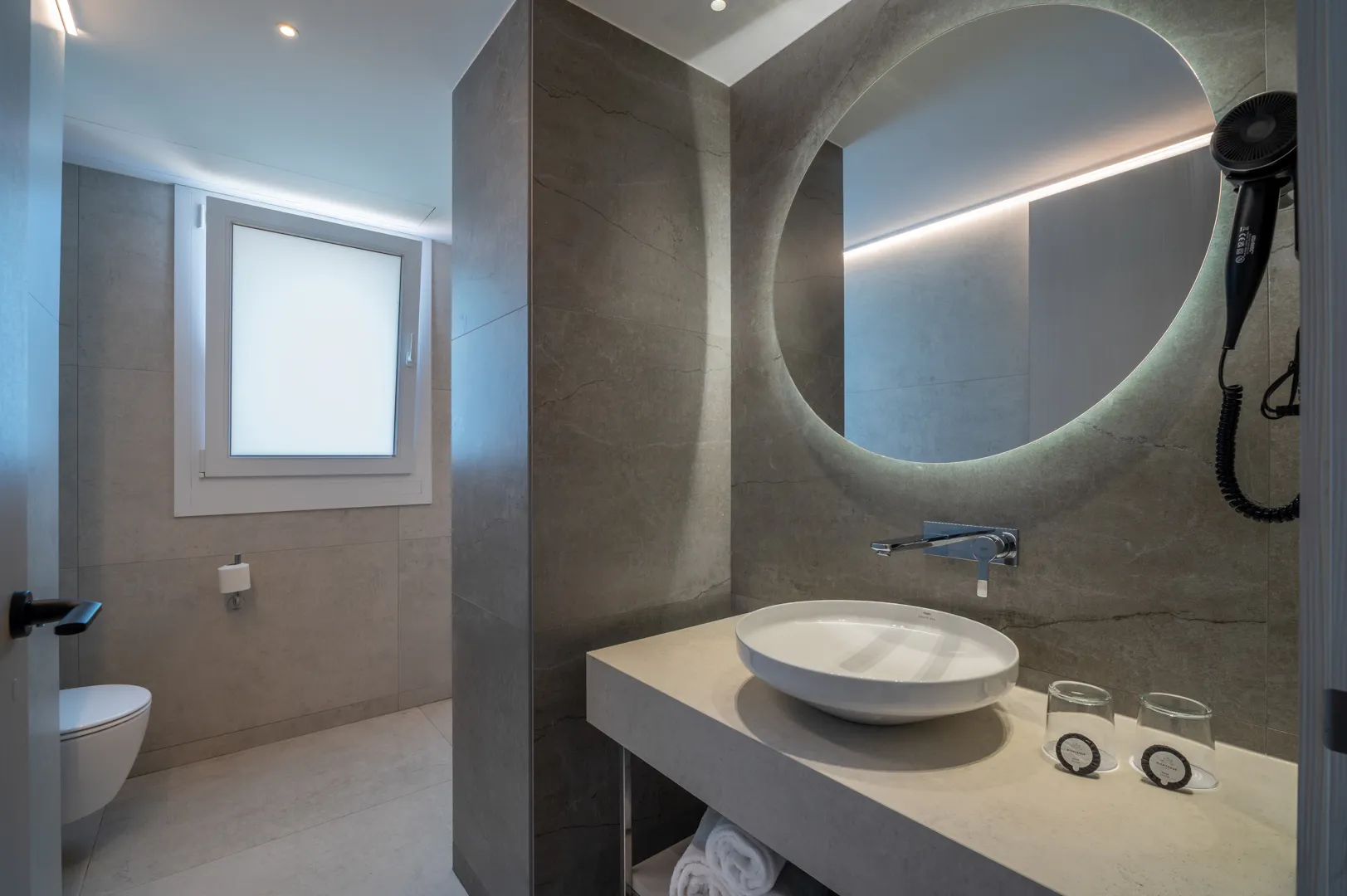 Baño moderno con inodoro, lavabo, espejo iluminado, secador y ventana. Costa Encantada Suites & Resort
