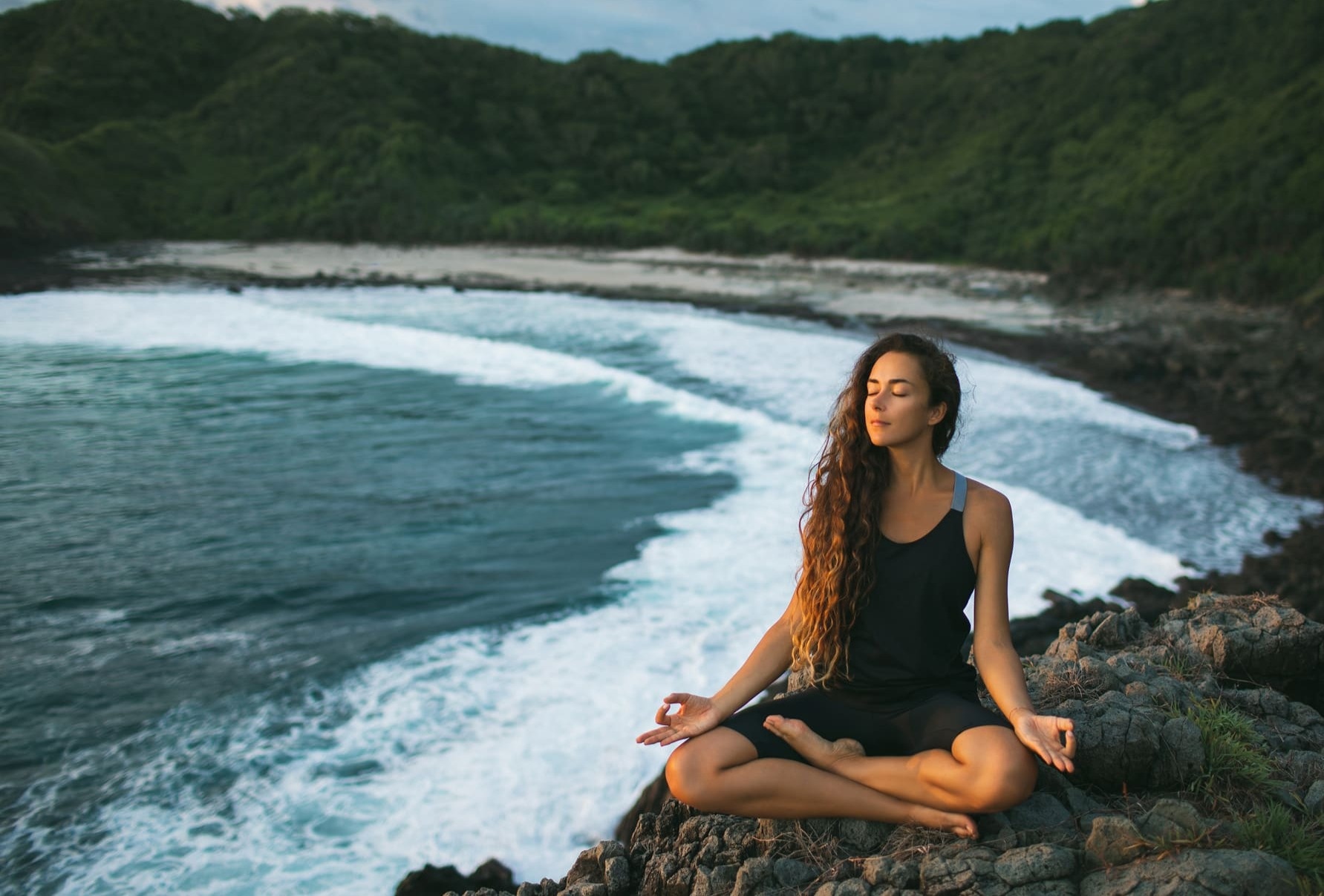 a woman sits on a rock near the ocean with her eyes closed