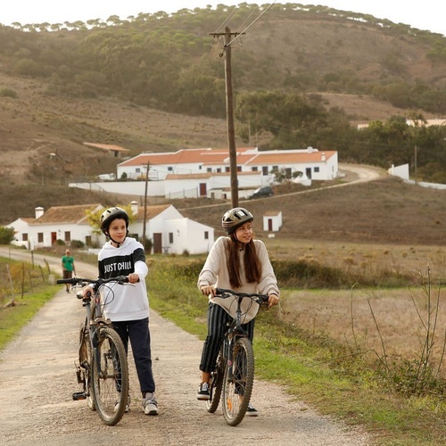 dos chicas andan en bicicleta por un camino de tierra con un burro en el fondo