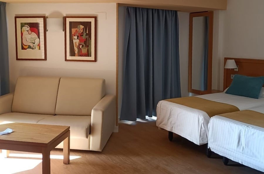 A bright room with light wood flooring features a beige sofa and coffee table, two framed artworks on the wall, and two single beds with blue curtains in the background.