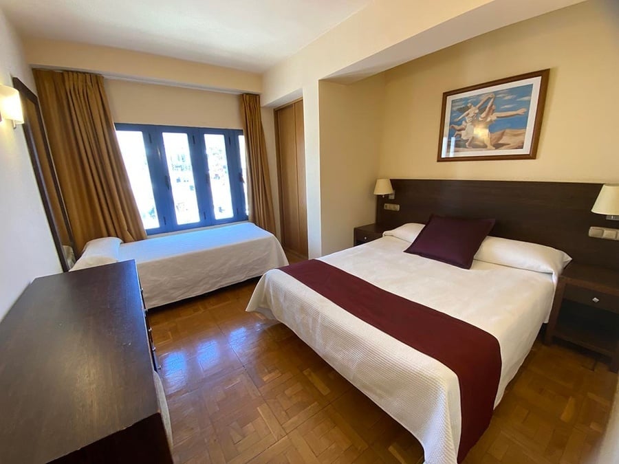 Dormitorio luminoso con una cama doble y una individual, una ventana con marcos azules y cortinas marrones.