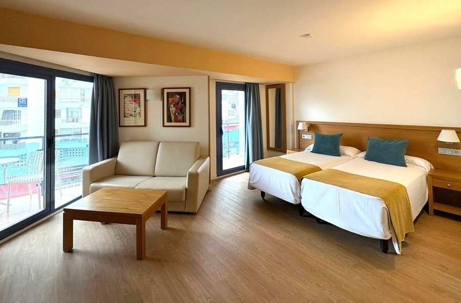 A bright and modern room, possibly a hotel room or studio apartment, features two twin beds with teal pillows, a beige sofa, a wooden coffee table, and large glass doors opening to a balcony with an urban view.