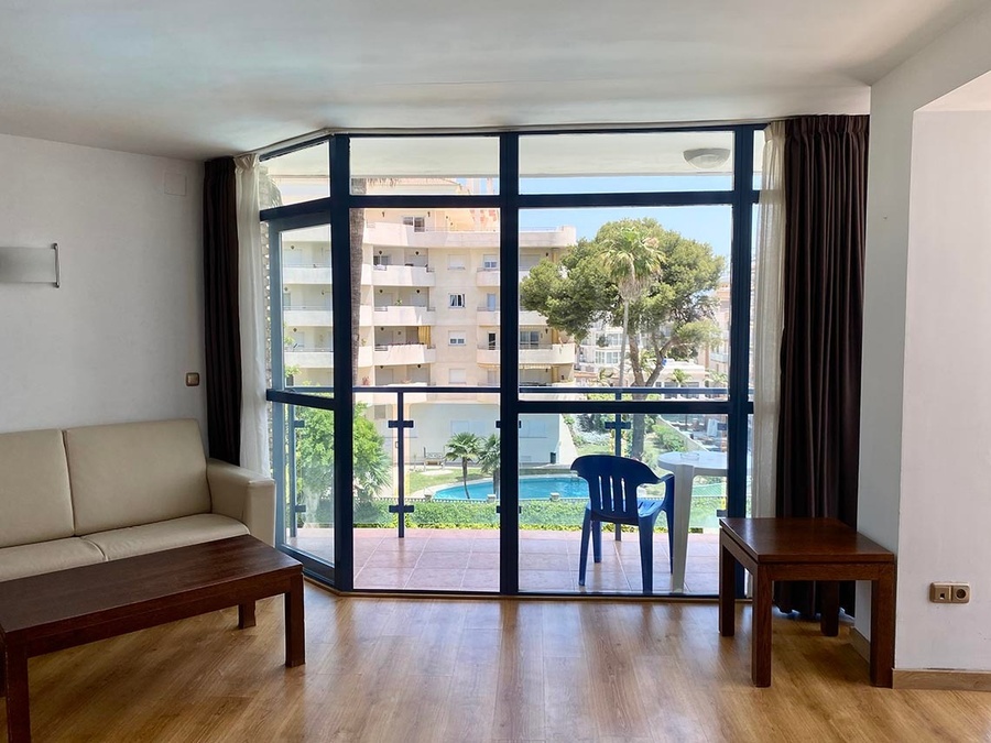 Una sala de estar con un sofá y mesas de madera ofrece una vista a través de un gran ventanal hacia una piscina, palmeras y edificios de apartamentos.