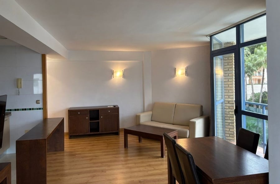 A bright open-plan living and dining area features wooden flooring, neutral walls with accent lighting, dark wood furniture, a light-colored sofa, and a large window revealing palm trees and buildings outside.