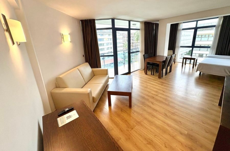 A bright, open-plan studio apartment features a living area with a cream sofa and coffee table, a dining area, and a bed, all against a backdrop of large windows with dark curtains providing an urban view.