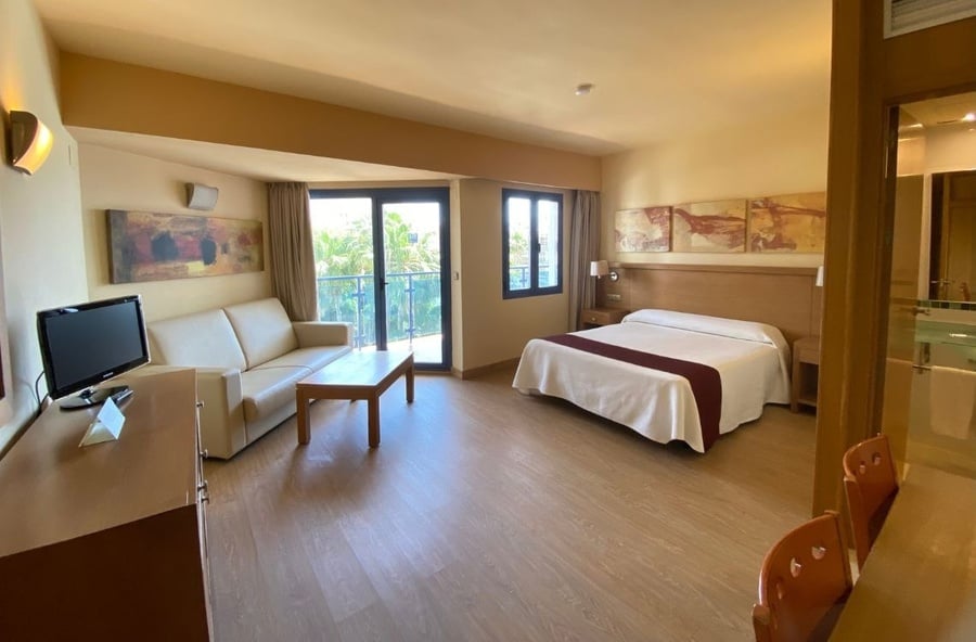 This image captures a bright and spacious hotel room featuring a bed, a sofa with a coffee table, a television, and a balcony overlooking lush greenery, all set on a light wood floor.
