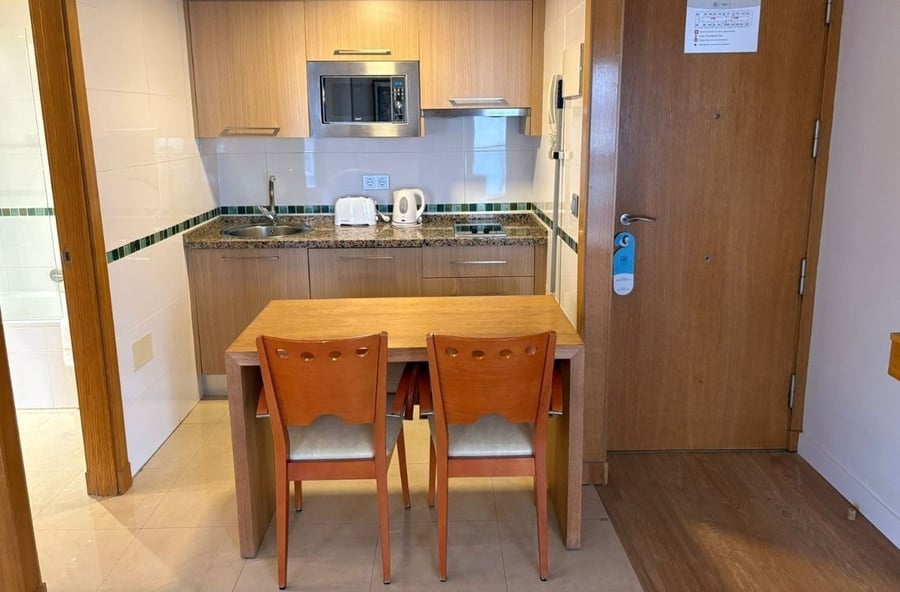 A compact kitchen area featuring light wooden cabinets, a granite countertop with various appliances, and a dining table with two chairs is visible, flanked by a wooden door on the right and a partial view of a bathroom entrance on the left.