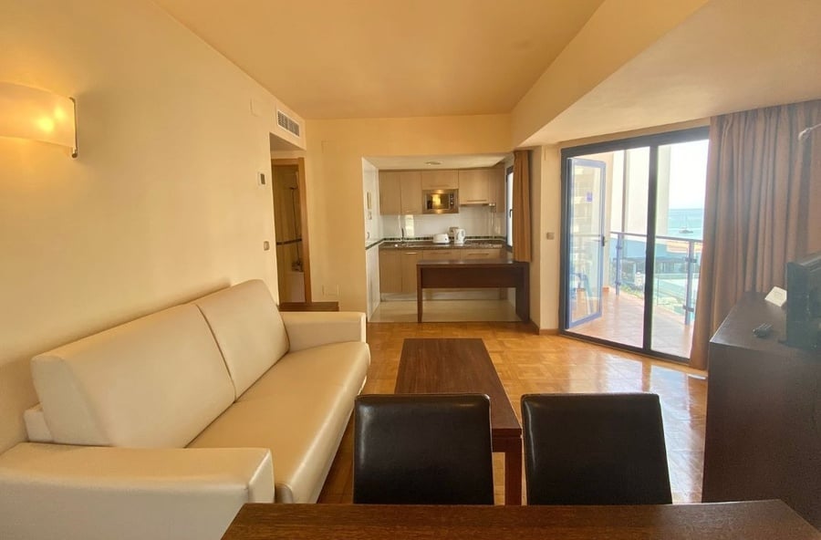 This image shows a bright, open-plan living area with a cream-colored sofa, dining table, and a compact kitchen in the background, all leading to a balcony with an ocean view.