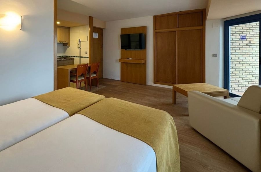 This image captures a bright studio apartment featuring two single beds, a small kitchen and dining area, a wall-mounted TV, and a living space with a sofa and coffee table.