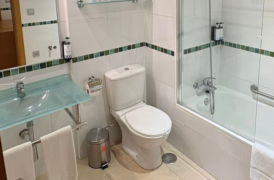 A clean and modern bathroom features white tiled walls with a green and blue accent strip, a glass sink with white towels, a toilet, and a bathtub with a glass shower screen.