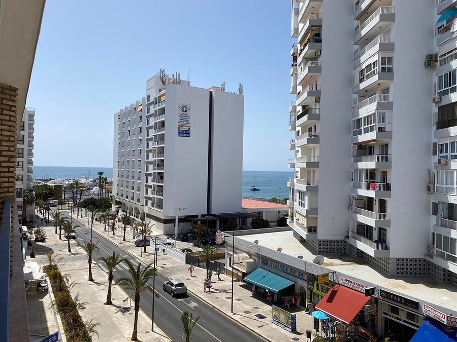 Vista panorámica de una calle costera flanqueada por altos edificios y palmeras, con tiendas a pie de calle y el mar Mediterráneo al fondo.