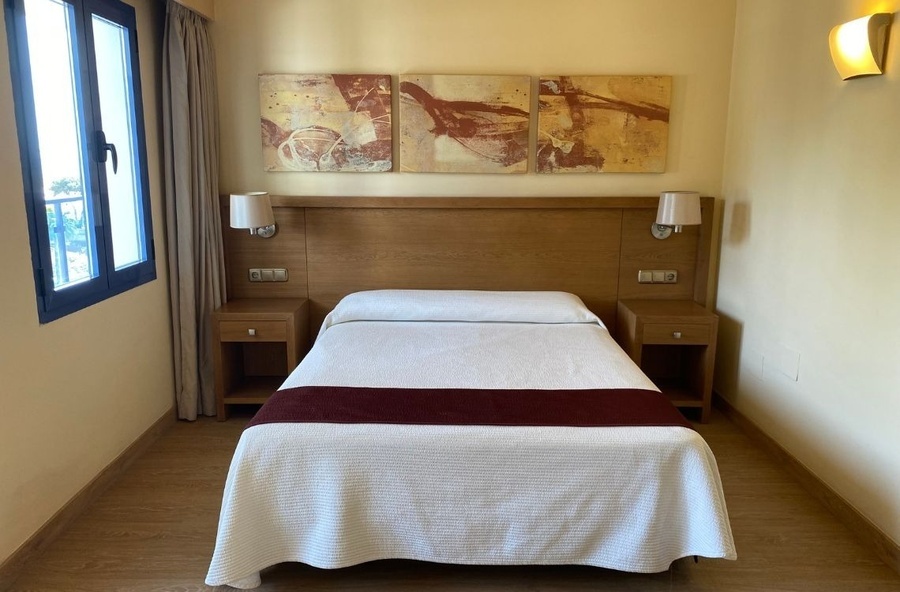 A neat and simple hotel-style bedroom features a bed with a wooden headboard, bedside tables with lamps, abstract paintings, a window, and wooden flooring.