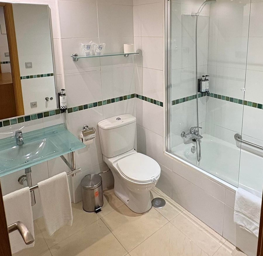 Este es un cuarto de baño luminoso con azulejos blancos y una franja decorativa verde, equipado con inodoro, lavabo de cristal, espejo y una bañera con ducha y mampara de cristal.