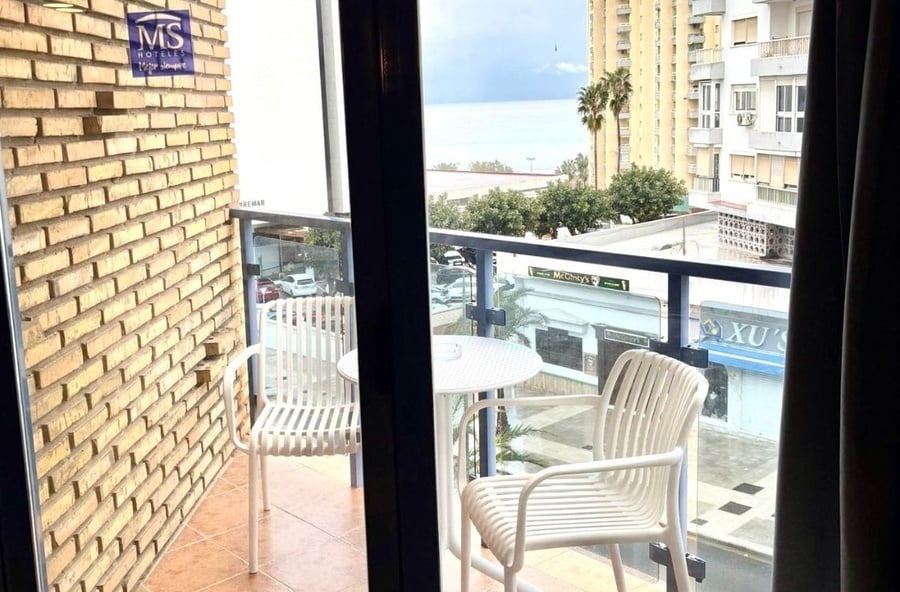 A small balcony with a table and two chairs offers a view of the sea, palm trees, and businesses below.