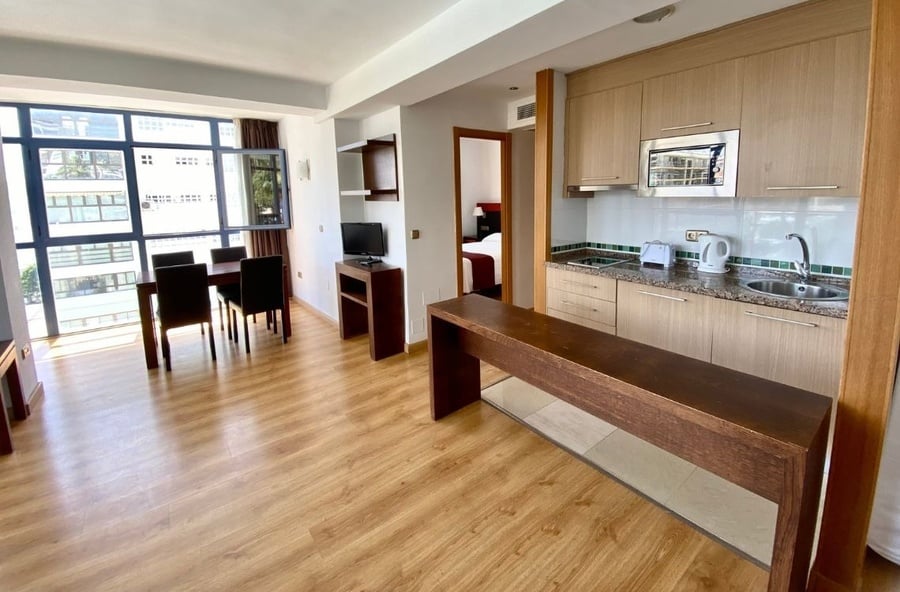 This image captures a bright, open-plan apartment featuring a dining area with a dark wooden table, a compact kitchen with light cabinetry, and large windows overlooking city buildings, with a bedroom visible through an open doorway in the background.