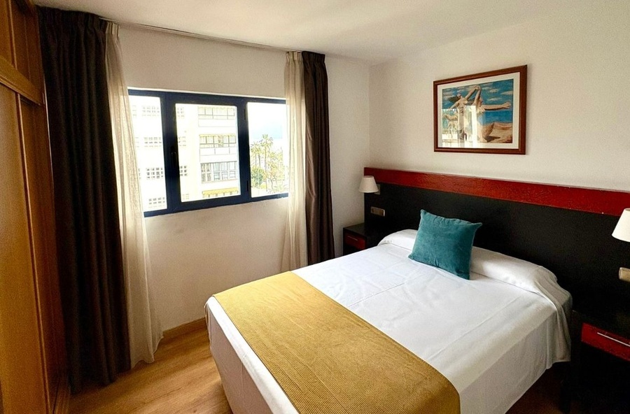 A cozy bedroom features a bed with a yellow runner and a teal pillow, a dark headboard with red trim, a framed artwork, and a window with dark curtains offering a view of buildings and palm trees.