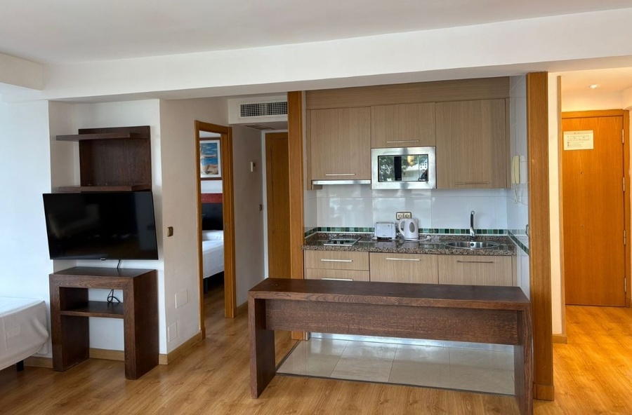 This image captures an open-plan studio apartment or hotel suite, showcasing a compact kitchen area with wooden cabinets and modern appliances, and a living space with a wall-mounted TV, with a bedroom visible through an open doorway.