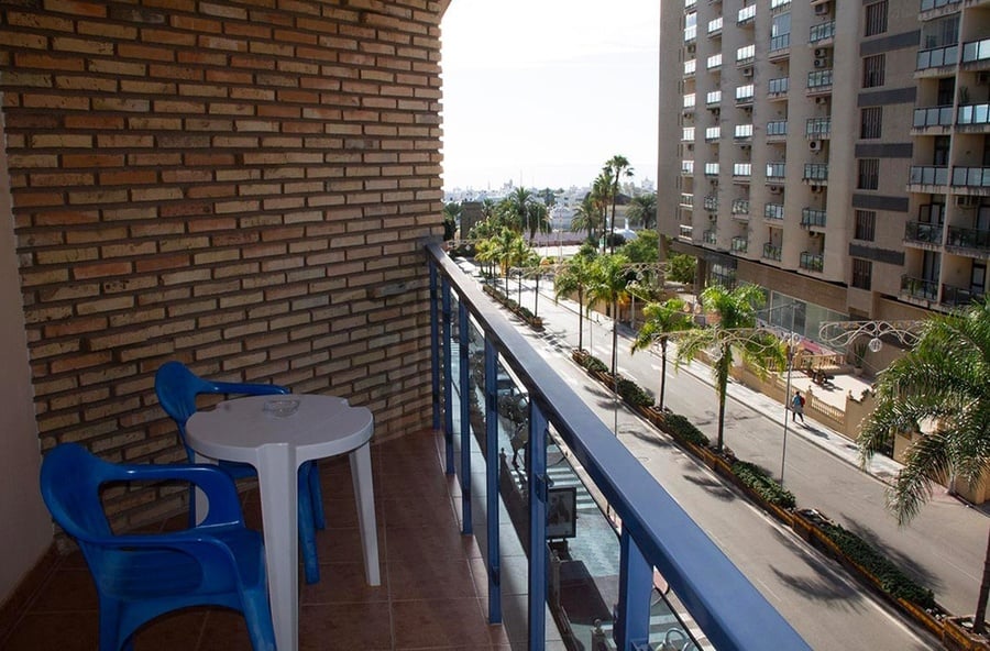 Un balcón con mesa y sillas azules ofrece vistas a una calle urbana flanqueada por palmeras y edificios altos.