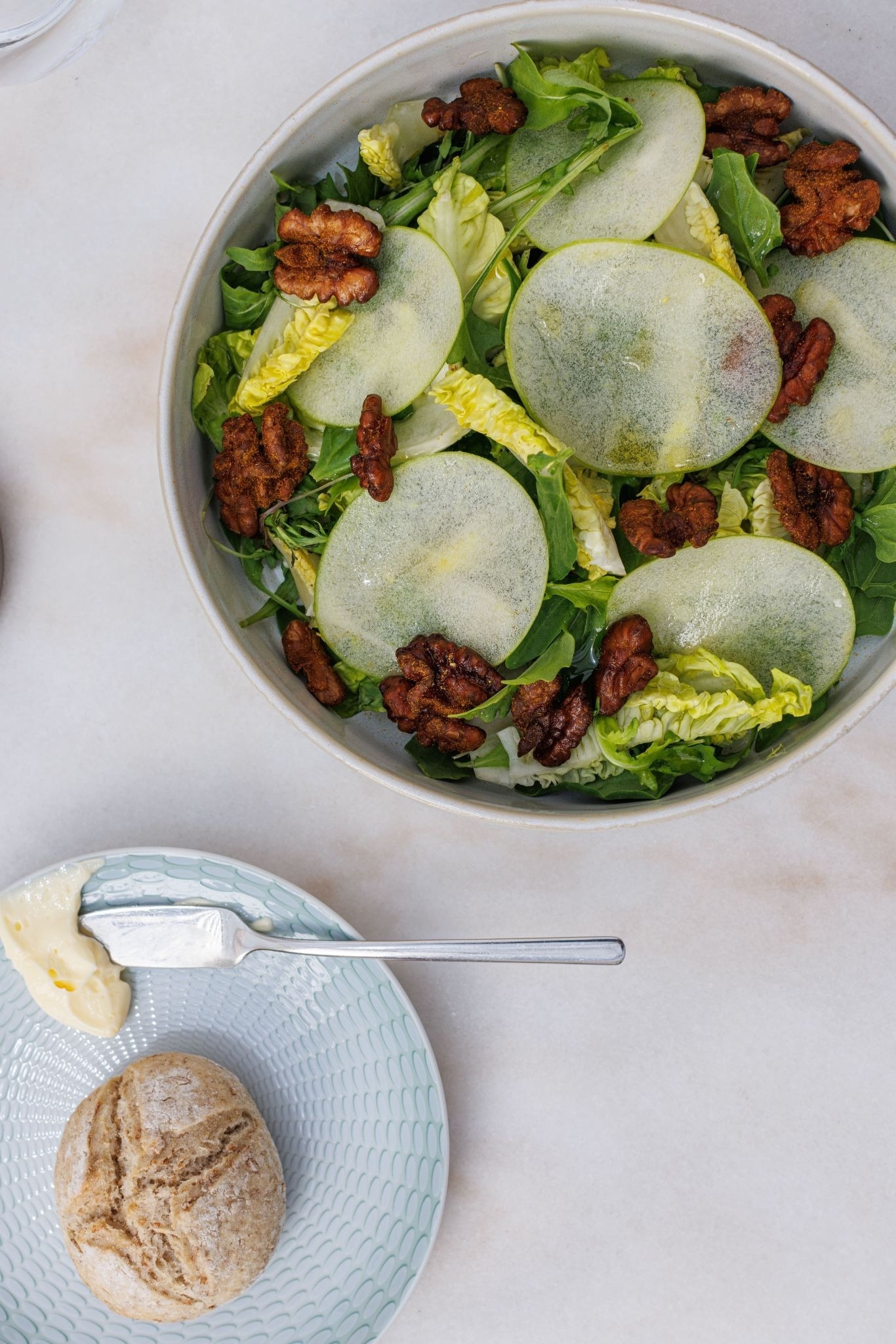 a bowl of salad next to a plate of butter and a roll of bread