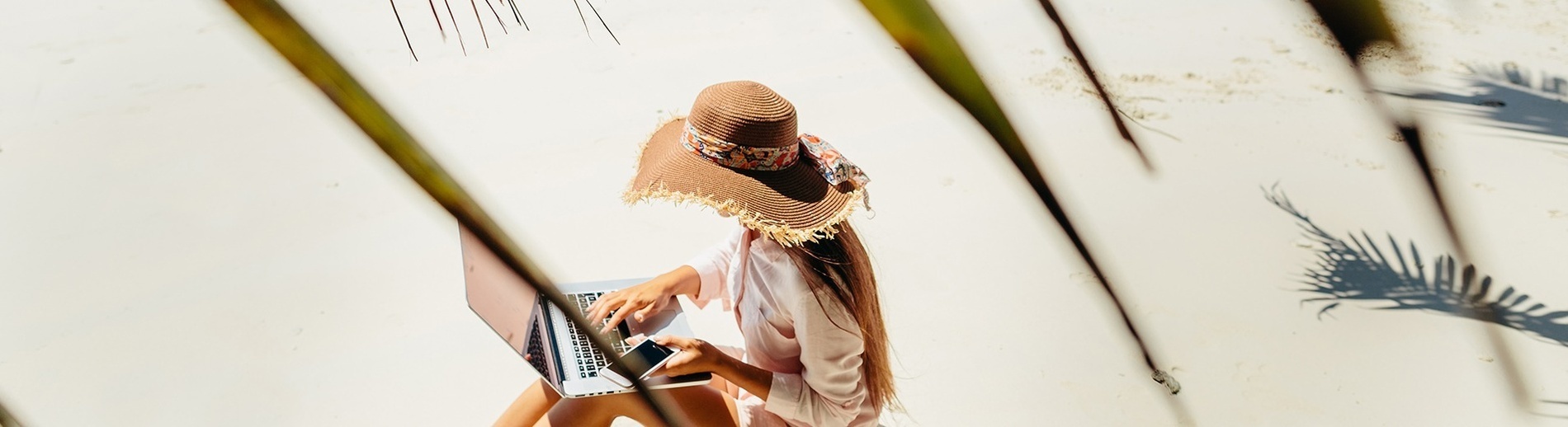 una mujer con un sombrero se sienta en la playa usando una computadora portátil