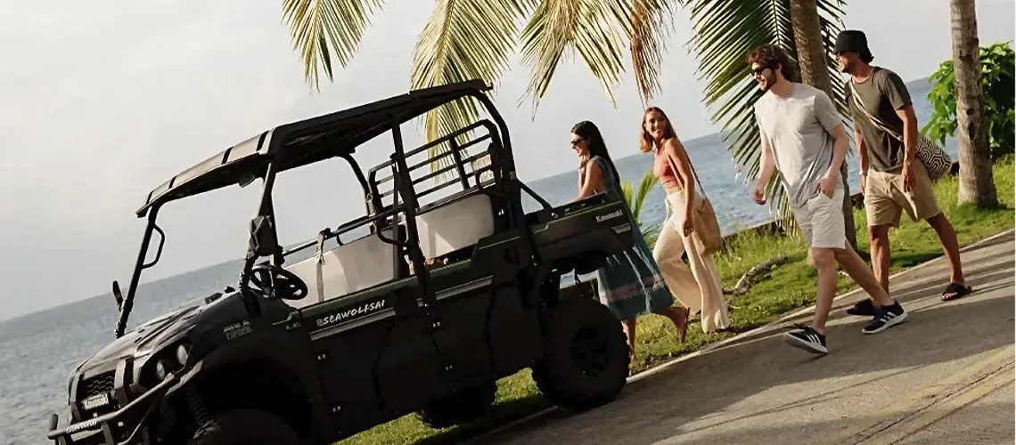 People walk past a Kawasaki Nattivo Collection Hotel vehicle near the ocean and palm trees on a sunny day.