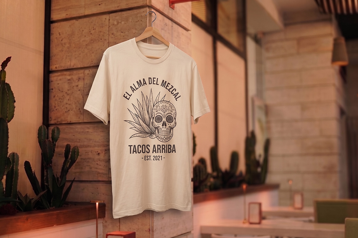 Camiseta com estampa de caveira e agave 'El Alma del Mezcal Tacos Arriba Nattivo Collection Hotel' em um ambiente rústico.