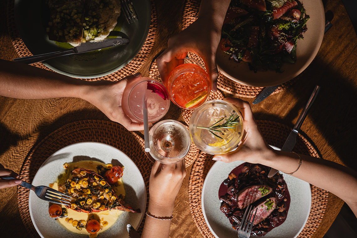 Cena gourmet con bebidas en mesa de madera, incluyendo langosta, carne y ensalada, perfecta para tu Nattivo Collection Hotel.