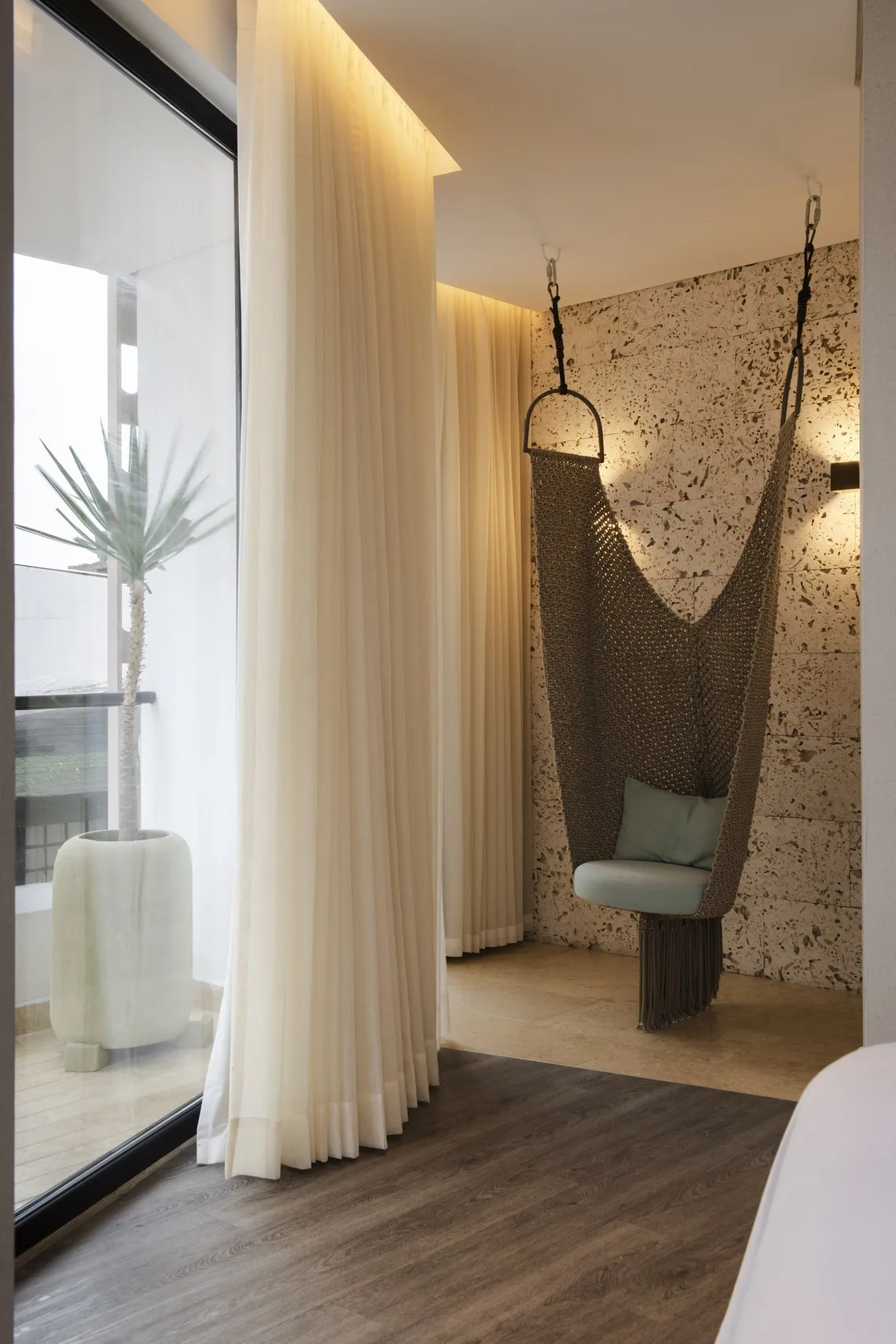 Cadeira de rede suspensa com almofada azul em quarto de hotel, perto de cortinas e janela com planta. Nattivo Collection Hotel