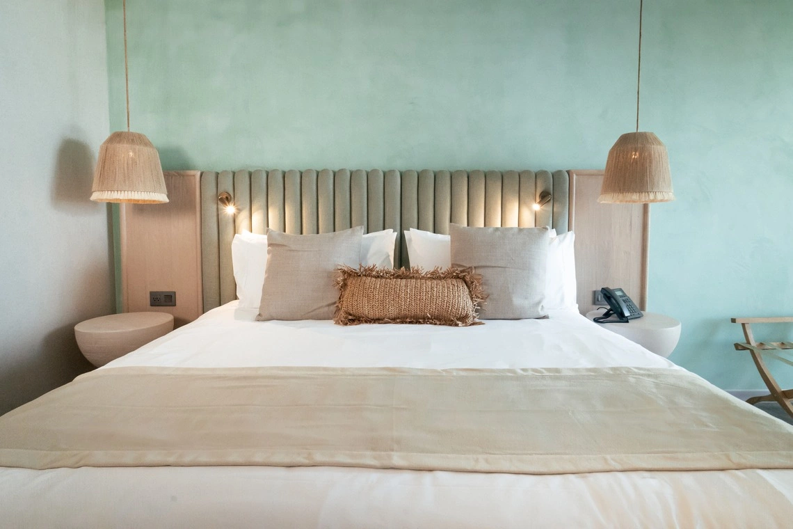 Cama de hotel confortável com almofadas neutras e luminária pendente de vime, perfeita para Nattivo Collection Hotel.