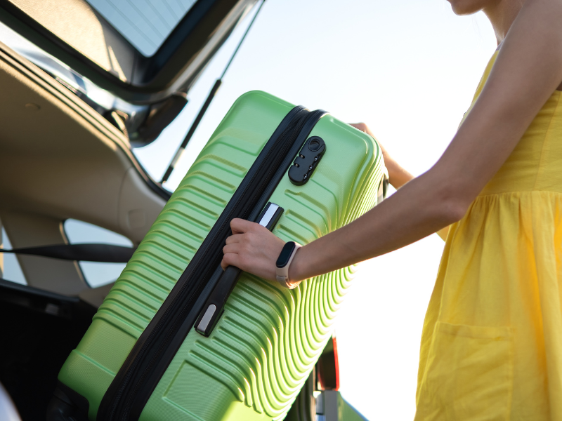 een vrouw in een gele jurk trekt een groene koffer uit de kofferbak van een auto