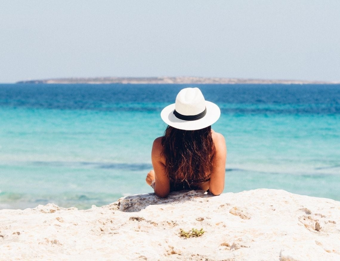 Una mujer con un sombrero blanco se sienta de espaldas, contemplando el mar azul turquesa desde una orilla rocosa.