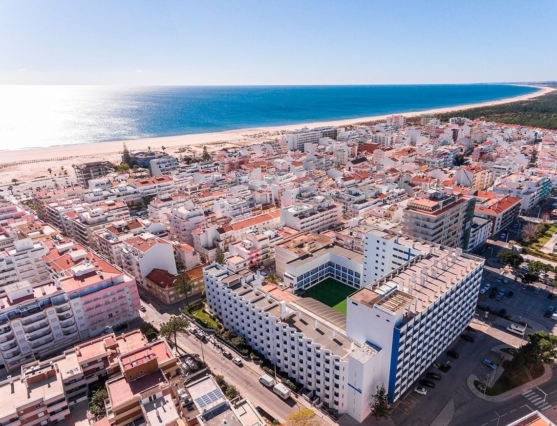 La imagen muestra una vista aérea de una ciudad costera densamente edificada, con una extensa playa y el mar azul bajo un cielo despejado.