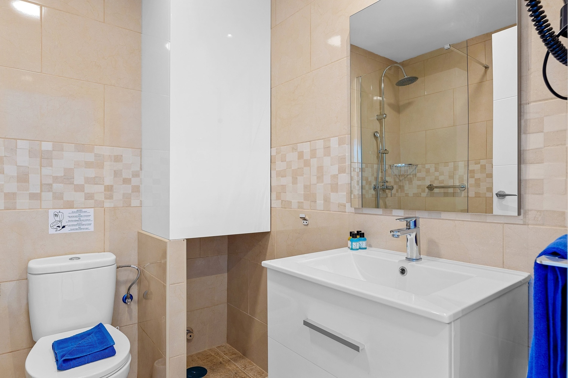 Une salle de bain moderne et lumineuse, carrelée de tons beiges avec une bande décorative, dotée de toilettes, d'un lavabo avec un meuble sous-vasque et d'un grand miroir reflétant une douche.