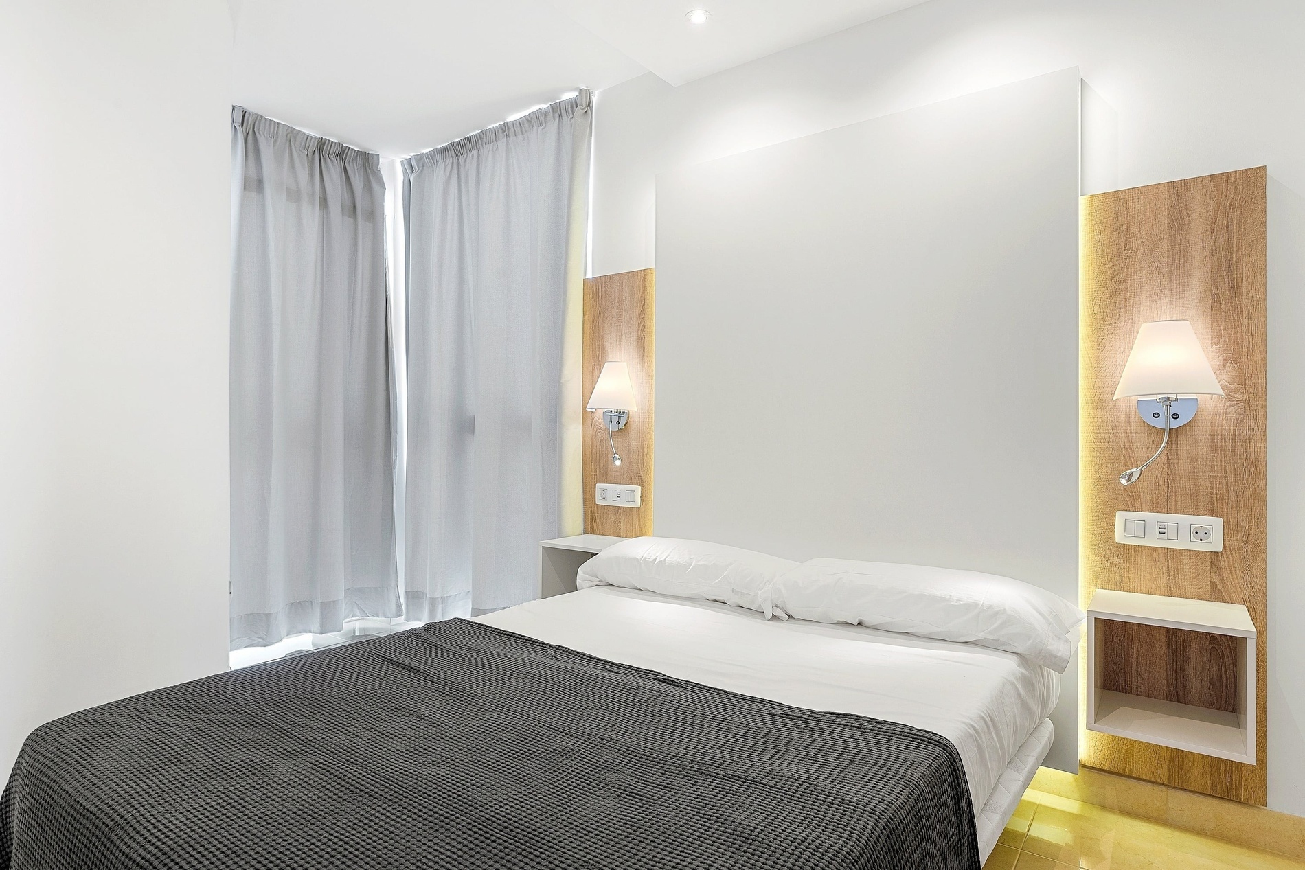 Une chambre moderne et lumineuse est présentée avec un lit fait, des rideaux gris, et un panneau de tête de lit en bois clair intégrant une applique murale, une liseuse, des prises électriques et un chevet.