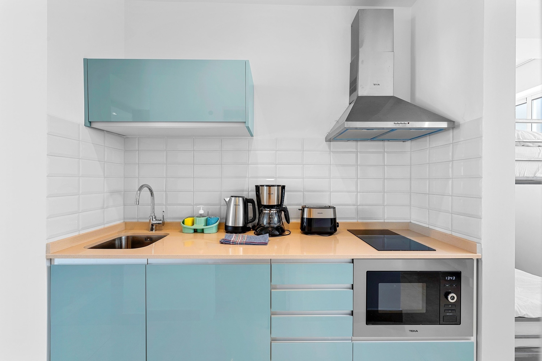 Une cuisine moderne et compacte est présentée avec des armoires inférieures et supérieures de couleur bleu canard, un carrelage blanc en métro, un plan de travail couleur sable et divers appareils électroménagers comme un four micro-ondes, une plaque de cuisson, une bouilloire et une cafetière.