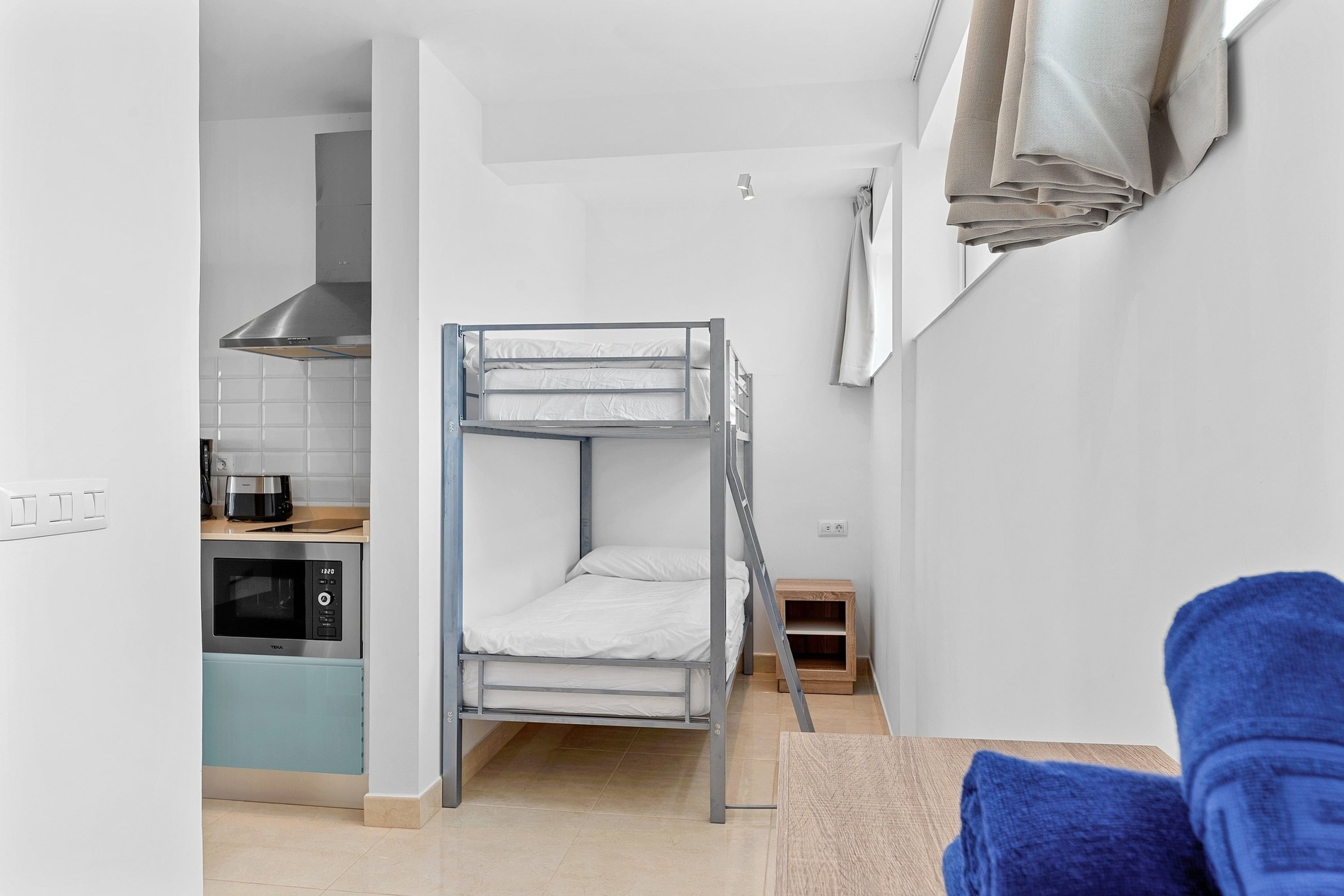 Une pièce lumineuse de style moderne abrite un lit superposé, un coin cuisine intégré et des serviettes bleues posées sur une table au premier plan.