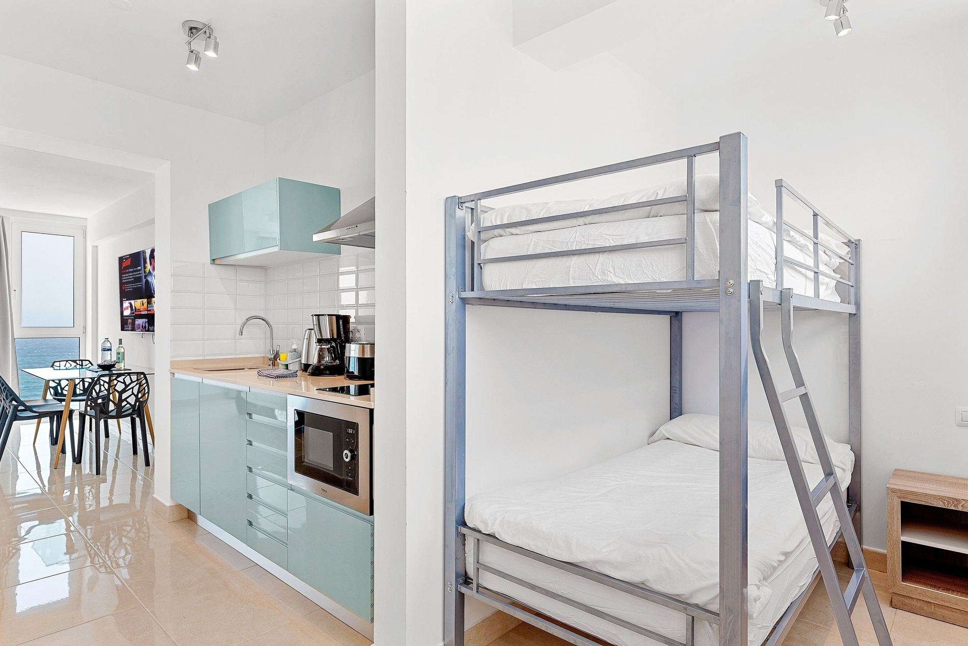 Cette image montre un intérieur moderne et lumineux comprenant une cuisine aux meubles bleu clair, un lit superposé et un coin repas offrant une vue imprenable sur la mer.