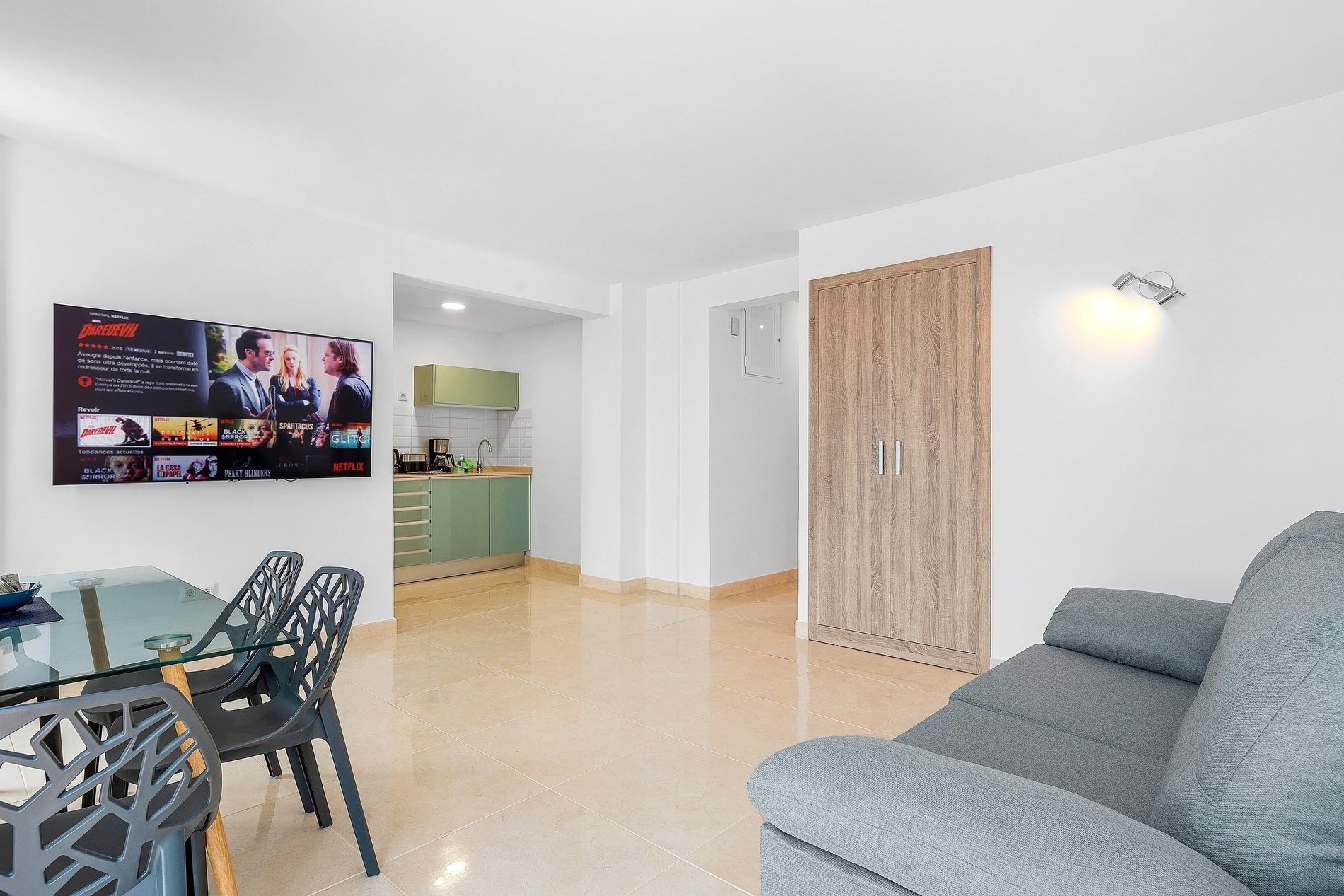 L'image présente un salon-salle à manger moderne et lumineux avec une télévision murale diffusant Netflix, une table en verre avec des chaises design, un canapé gris et une petite cuisine ouverte aux placards vert clair.